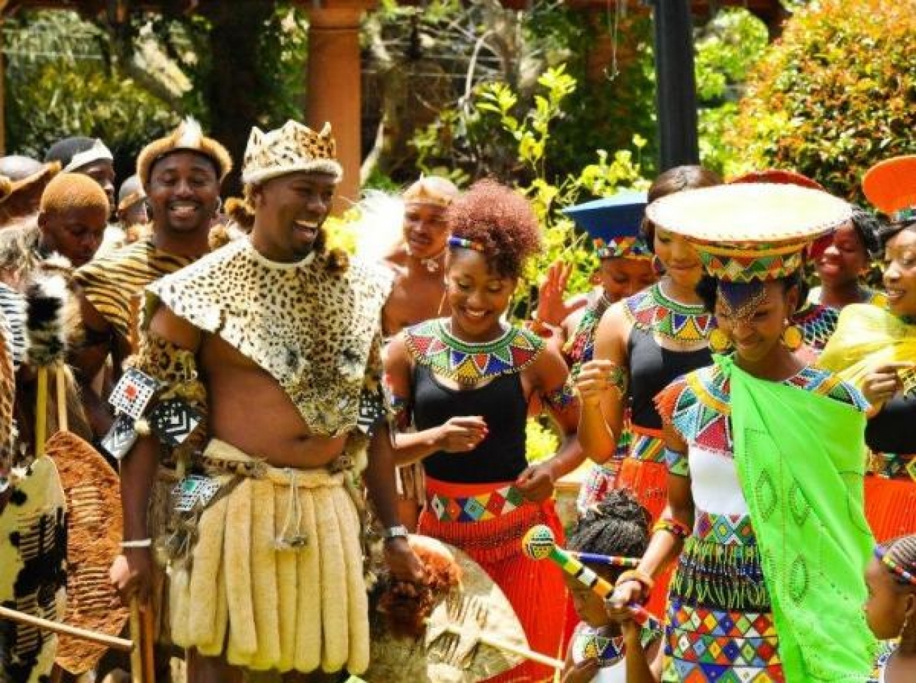 A group of people in traditional Zulu cultural attire laugh and dance against a green leafy background.