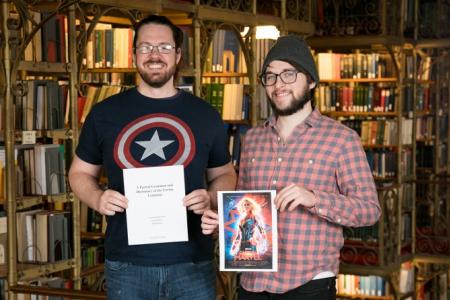 Graduate students Ryan Hearn, left, and Joseph Rhyne hold the grammar guide and dictionary for the Torfan language and a poster for "Captain Marvel."